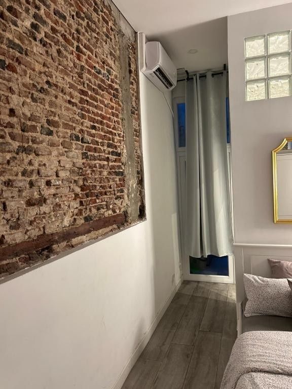 Pasillo estrecho con pared de ladrillo visto, paredes blancas, suelo gris y un pequeño rincón con ventana y cortina.