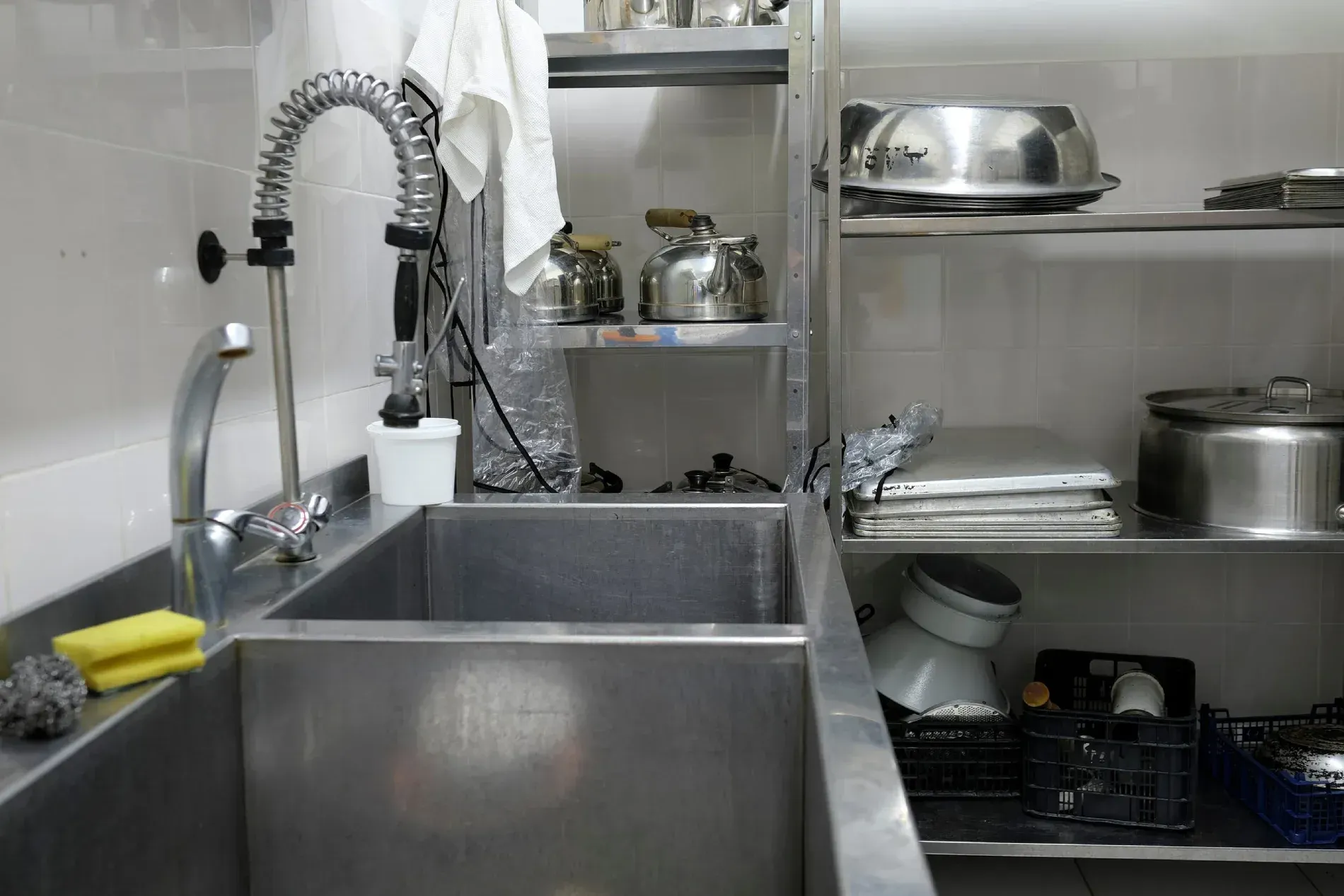 Fregadero de cocina comercial de acero inoxidable con rociador, platos, ollas y estantes.