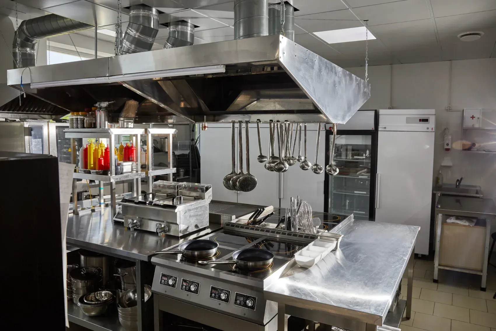Cocina comercial con estufa de acero inoxidable, campana extractora, utensilios colgantes y encimeras de preparación.