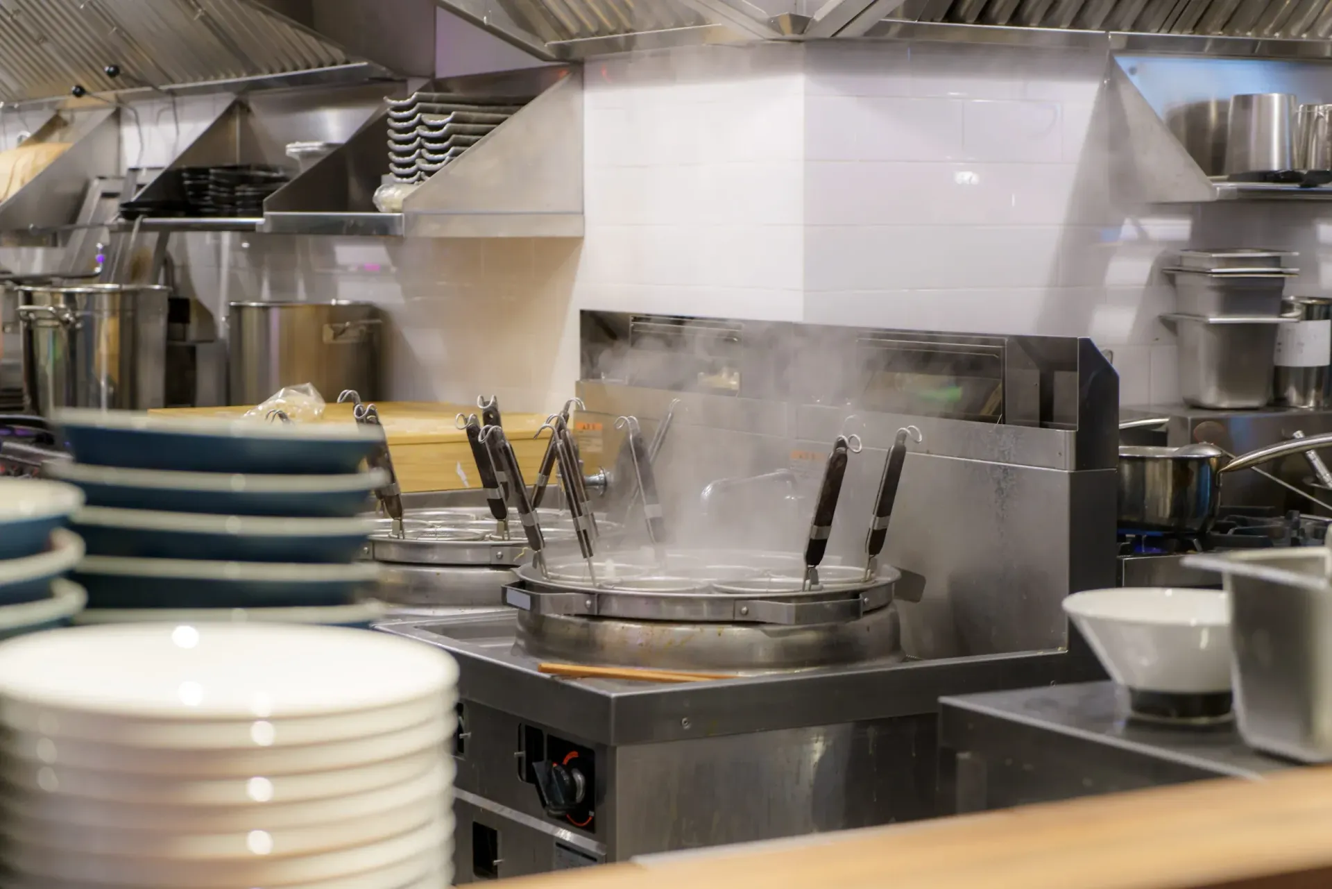 Cocina industrial con encimeras de acero inoxidable, platos apilados y equipo de cocina bajo luces brillantes.