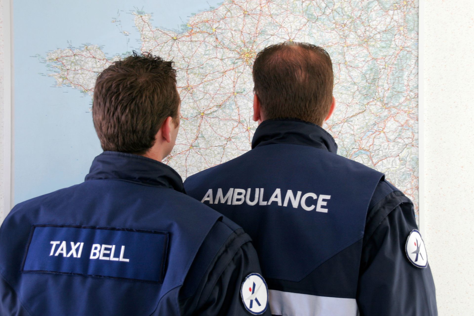 Deux employés en uniforme, portant les inscriptions TAXI BELL et AMBULANCE, étudient une carte murale.