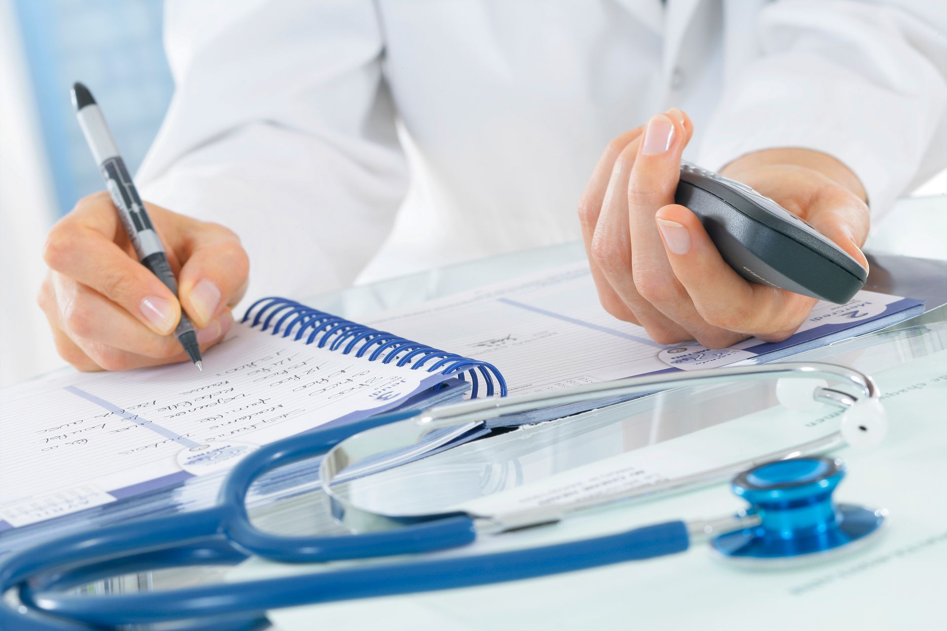 Un médecin écrivant sur un bloc-notes, un stéthoscope et une calculatrice posés sur un bureau.