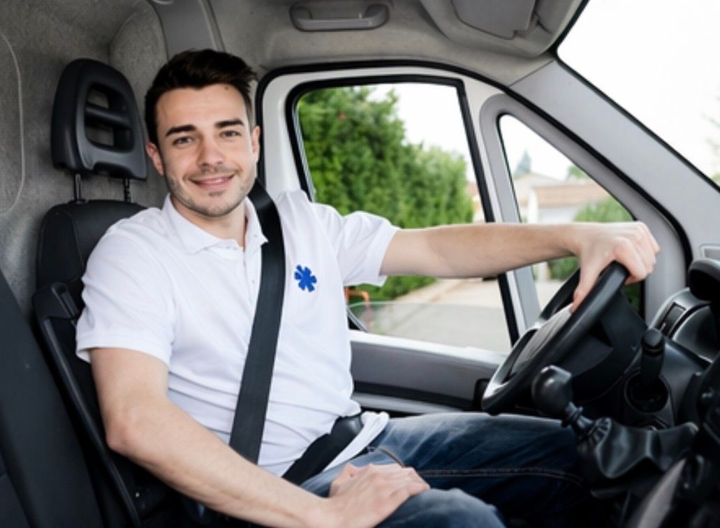 Un ambulancier souriant en chemise blanche est assis dans une fourgonnette médicale, la main sur le volant.