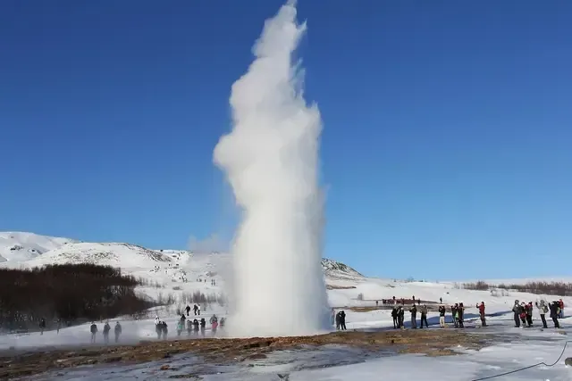 Un géiser entra en erupción, expulsando vapor hacia el cielo azul. La gente observa desde un paisaje nevado.