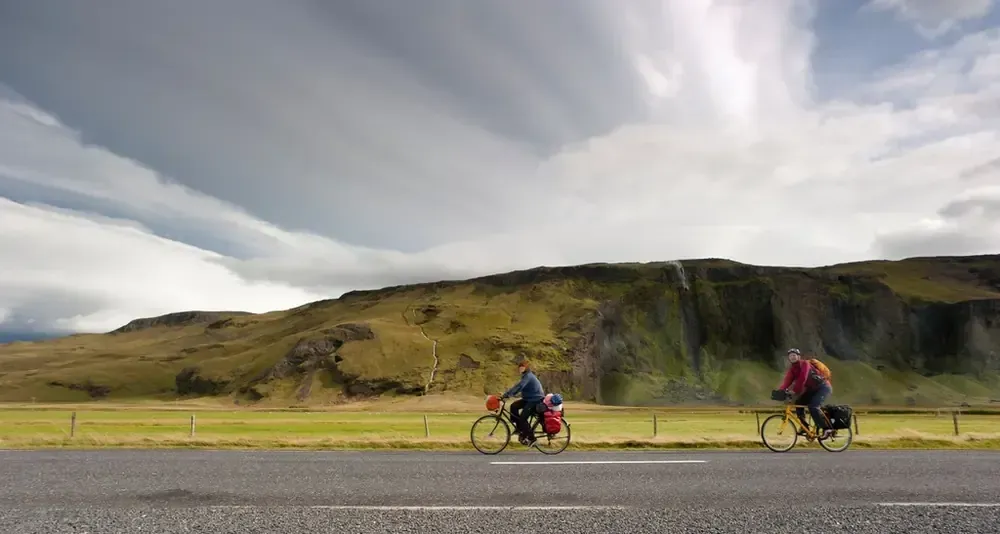 Dos ciclistas viajan por una carretera con un fondo de montañas bajo un cielo nublado.