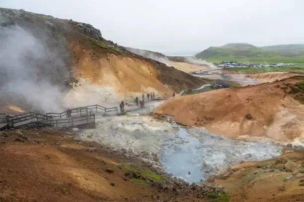 El vapor se eleva desde una zona geotérmica con pasarelas y algunas personas. Laderas de color marrón y canela.