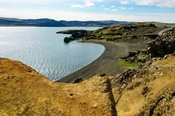 Una playa de arena negra curva se encuentra con un lago tranquilo bajo un cielo azul nublado.