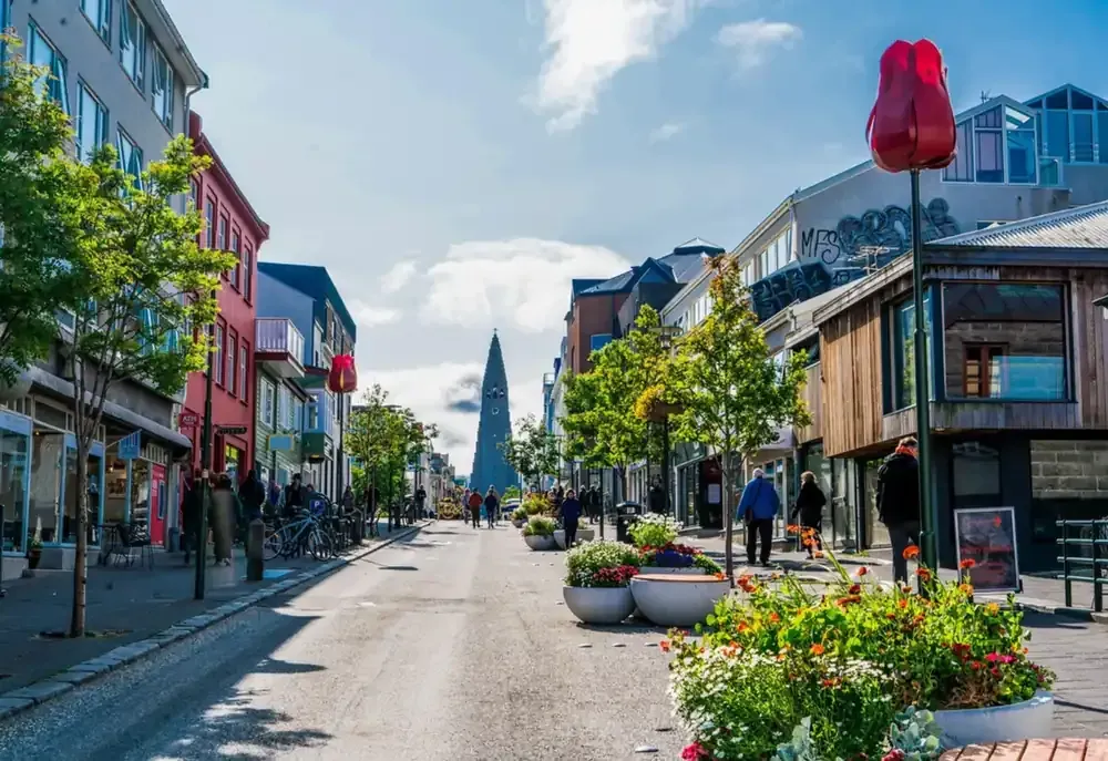 Escena callejera en Reykjavik, Islandia, con tiendas, gente, árboles y un monumento alto.