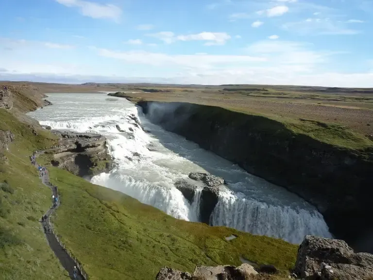Cascada cayendo por un acantilado hacia un río. Colinas cubiertas de hierba y cielo azul.