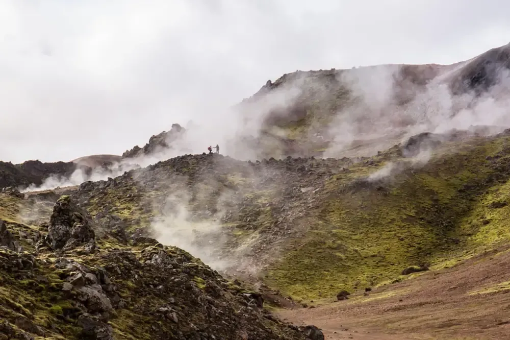 Terreno montañoso rocoso con vapor elevándose, algunas figuras en una cresta, cielo nublado.
