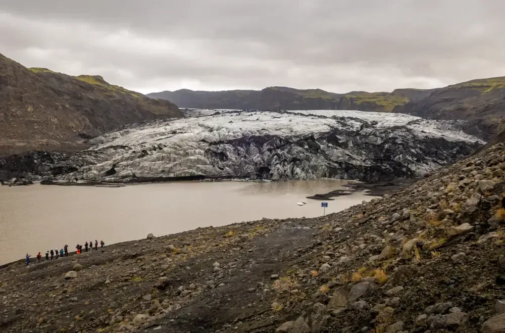 Glaciar con un fondo rocoso y oscuro, un lago y un cielo nublado. Hay gente cerca del agua.