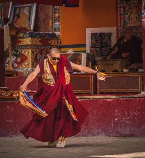 Un monje budista con túnicas rojas baila frente a un altar, sosteniendo un instrumento musical.