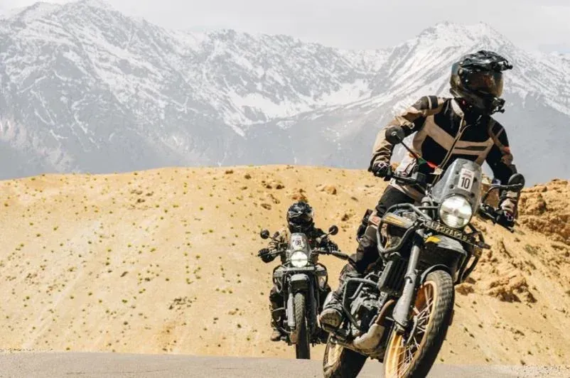 Dos motociclistas circulando por una carretera en un paisaje desértico con montañas al fondo.