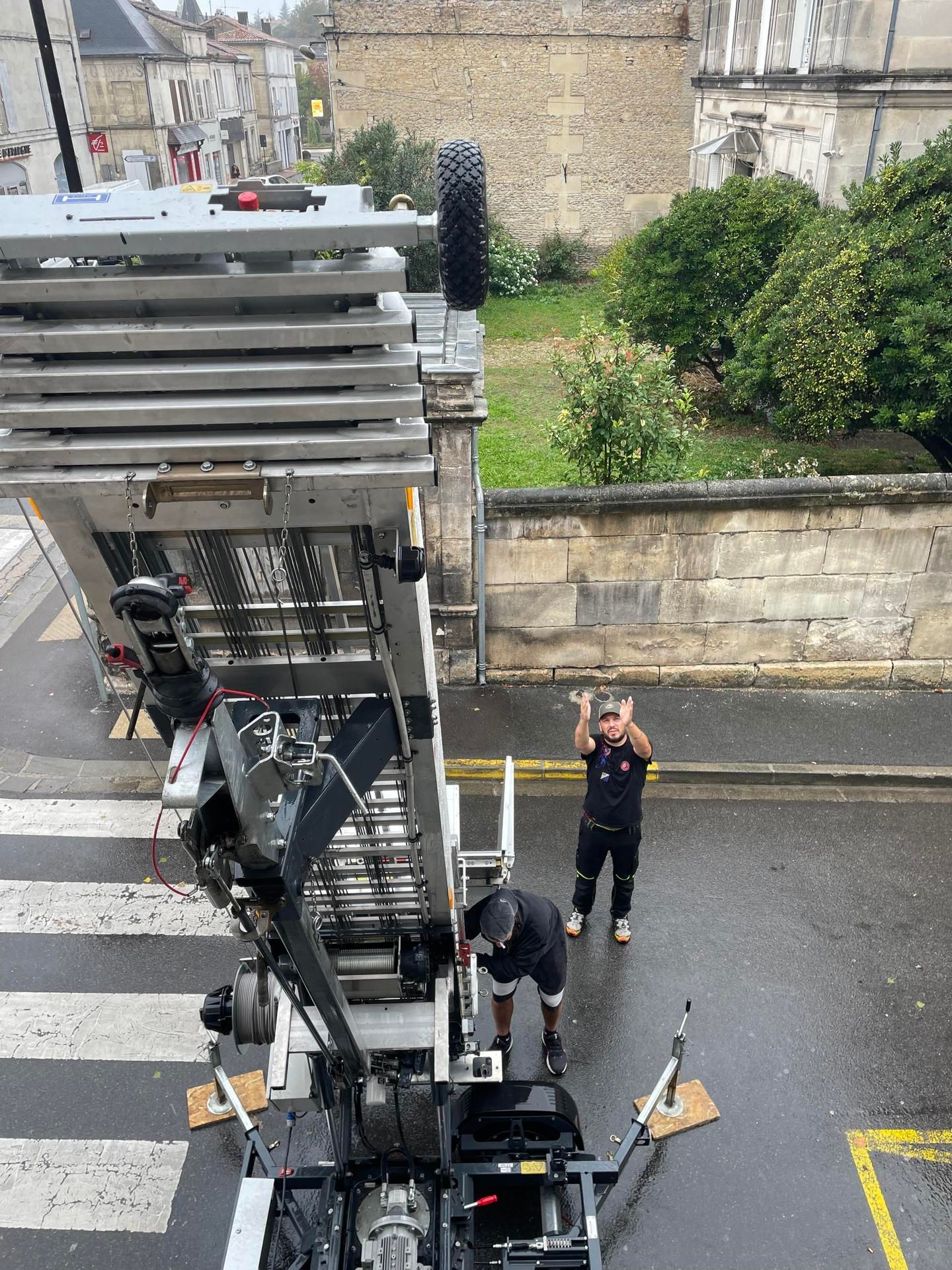 Deux déménageurs utilisent un élévateur pour transporter des objets. L'un dirige, l'autre conduit la machine, dans une rue pluvieuse.