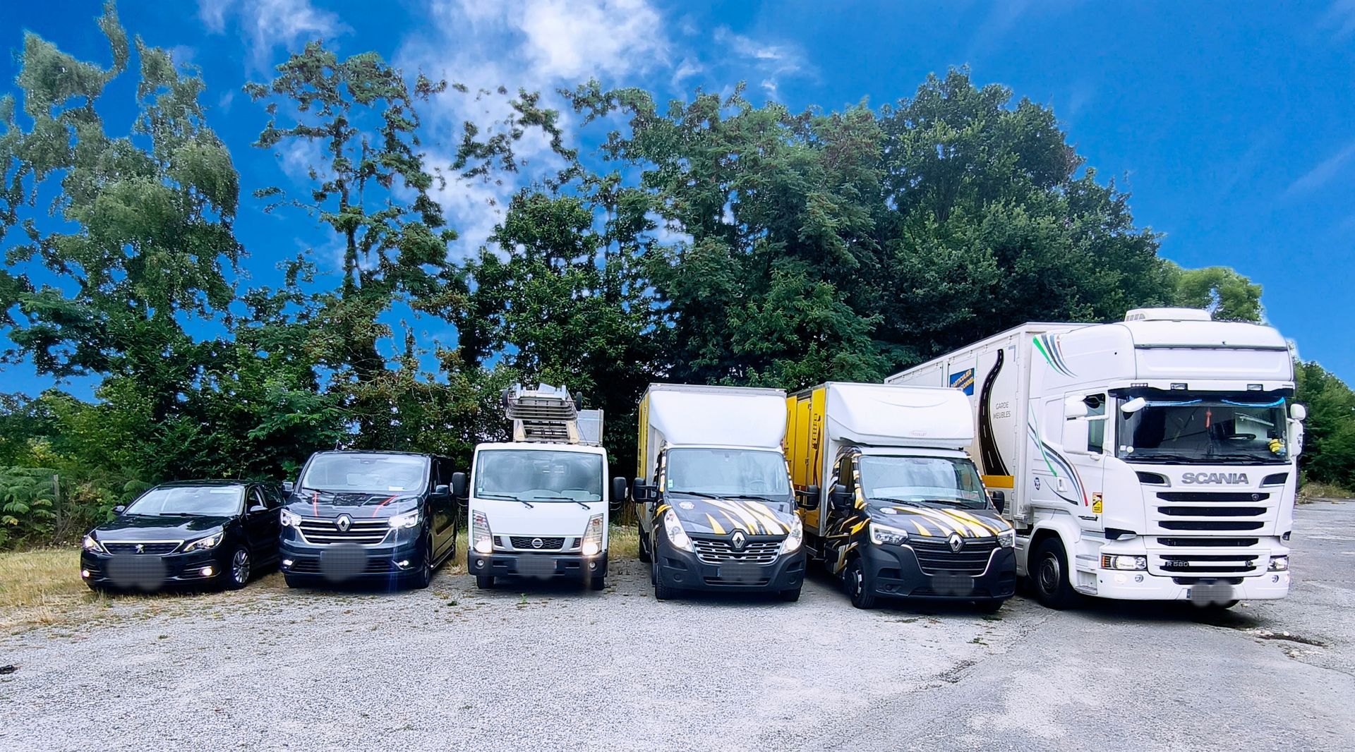 Véhicules de différentes tailles stationnés à l'extérieur : voiture, camionnettes et un semi-remorque sous un ciel bleu avec des arbres.