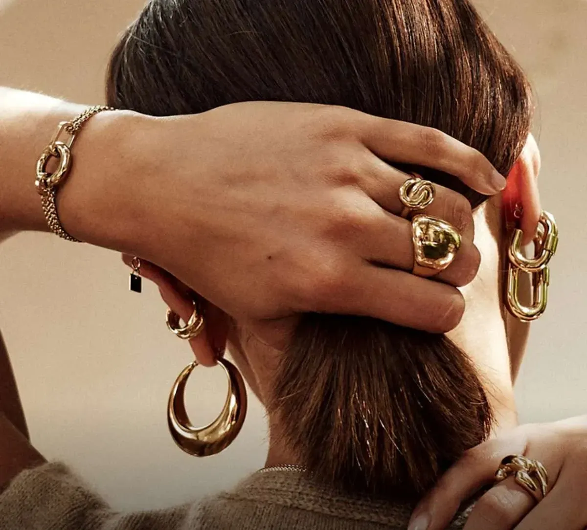 Les mains d'une femme, ornées de bijoux en or (bracelet, bagues et boucles d'oreilles), touchent ses cheveux.
