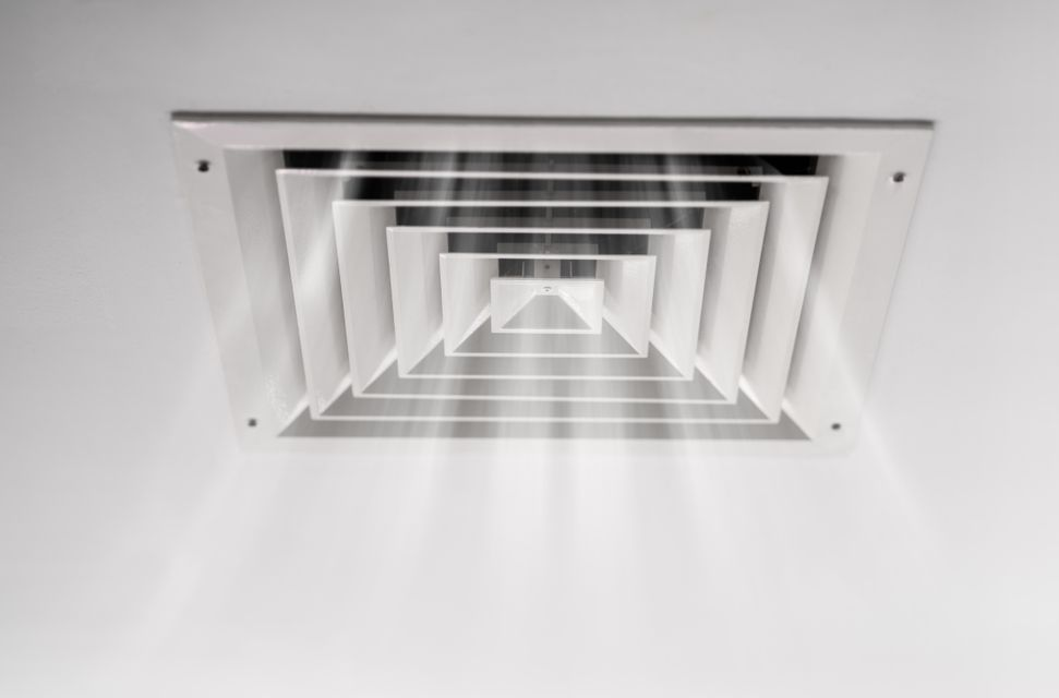 Une grille de ventilation de plafond, carrée et à persiennes, d'où émane une douce lumière en son centre.