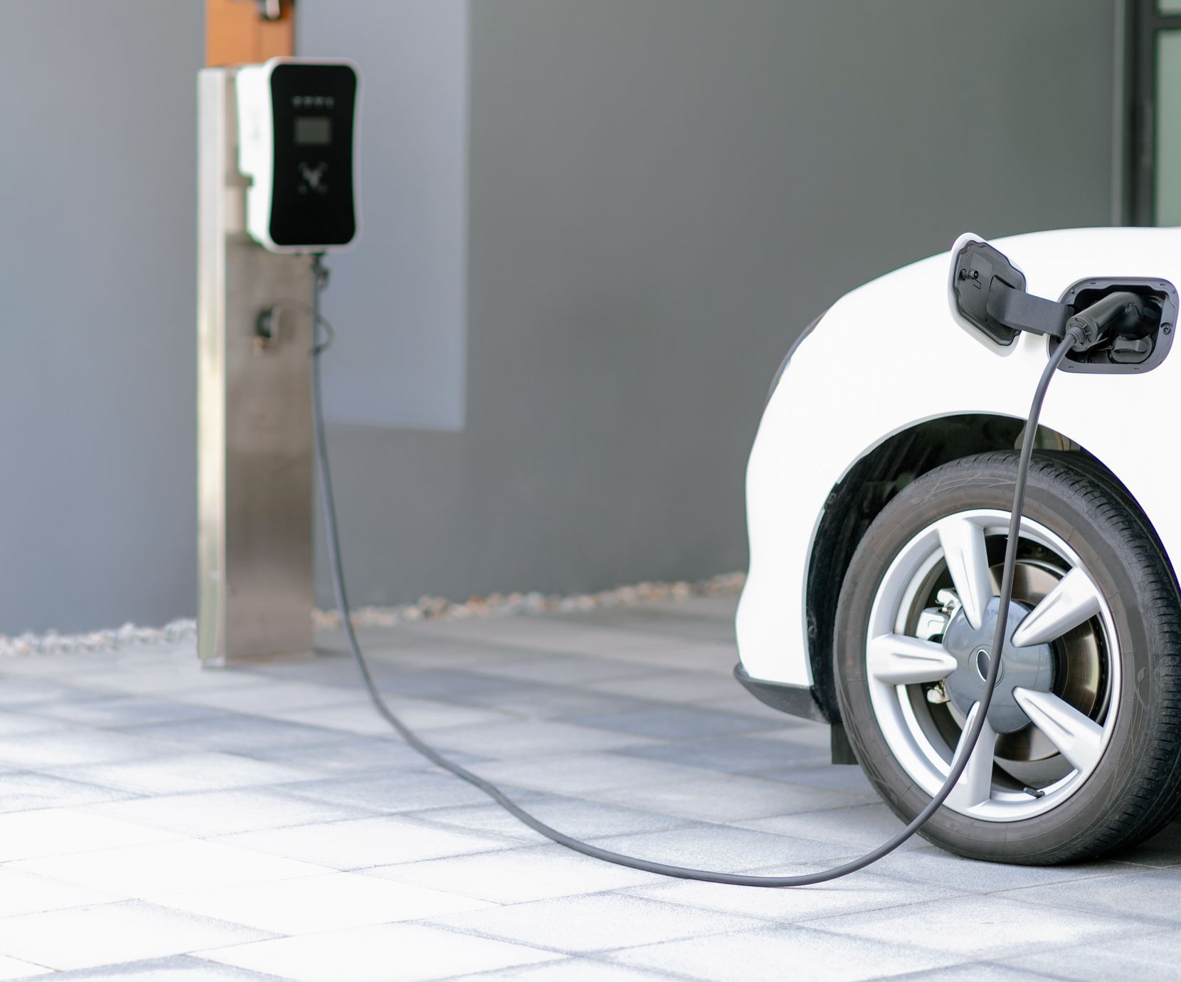 Une voiture électrique garée sur une allée pavée, en charge via un câble relié à une borne de recharge murale.