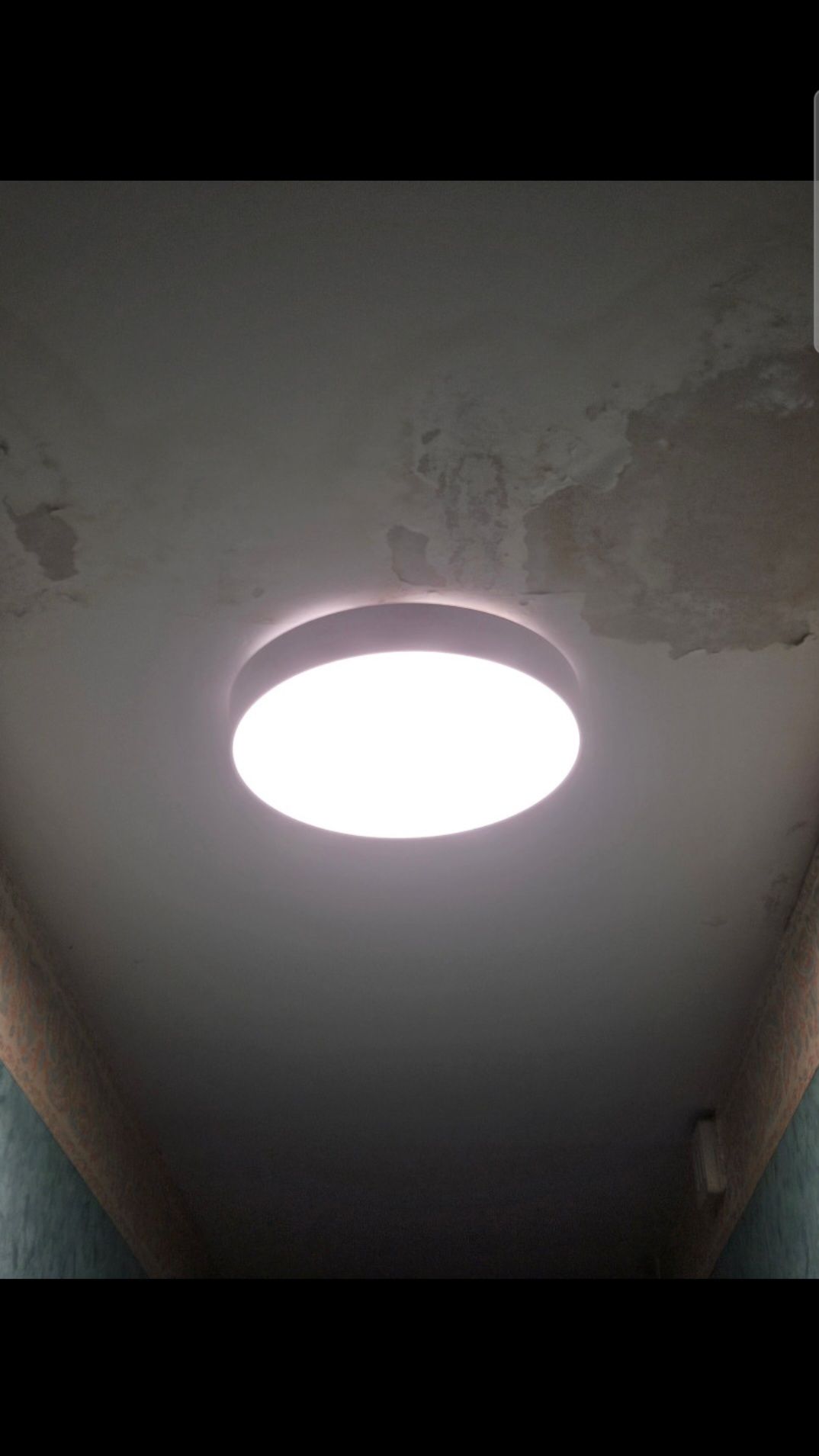 Un plafonnier circulaire encastré, lumineux, éclaire un plafond présentant quelques dommages superficiels visibles.