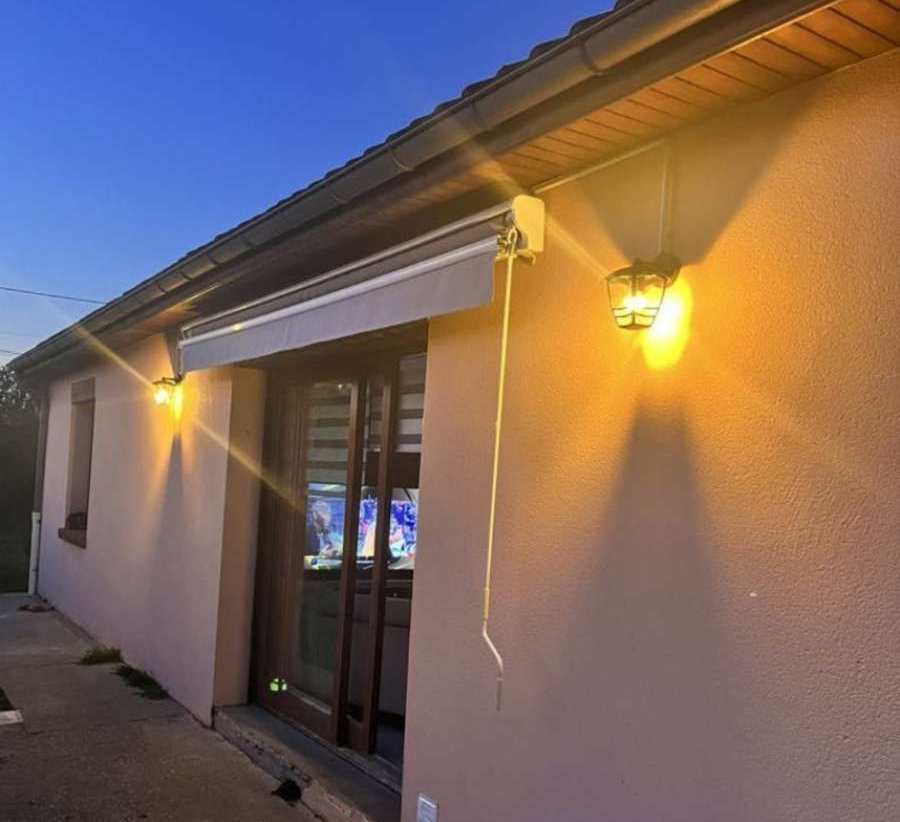 Un mur extérieur de maison au crépuscule, avec deux lanternes extérieures illuminées, un auvent de patio rétracté et une porte vitrée.