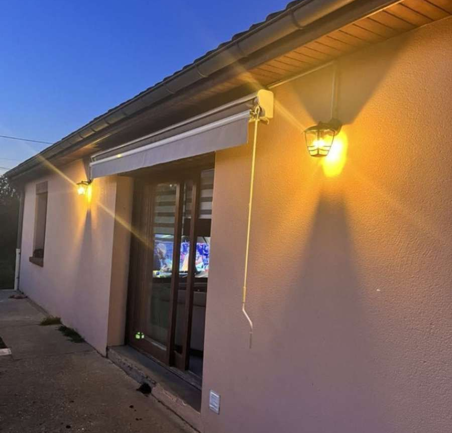 Mur extérieur d'une maison au crépuscule, avec deux appliques murales allumées et un store rétractable au-dessus d'une porte coulissante en verre.