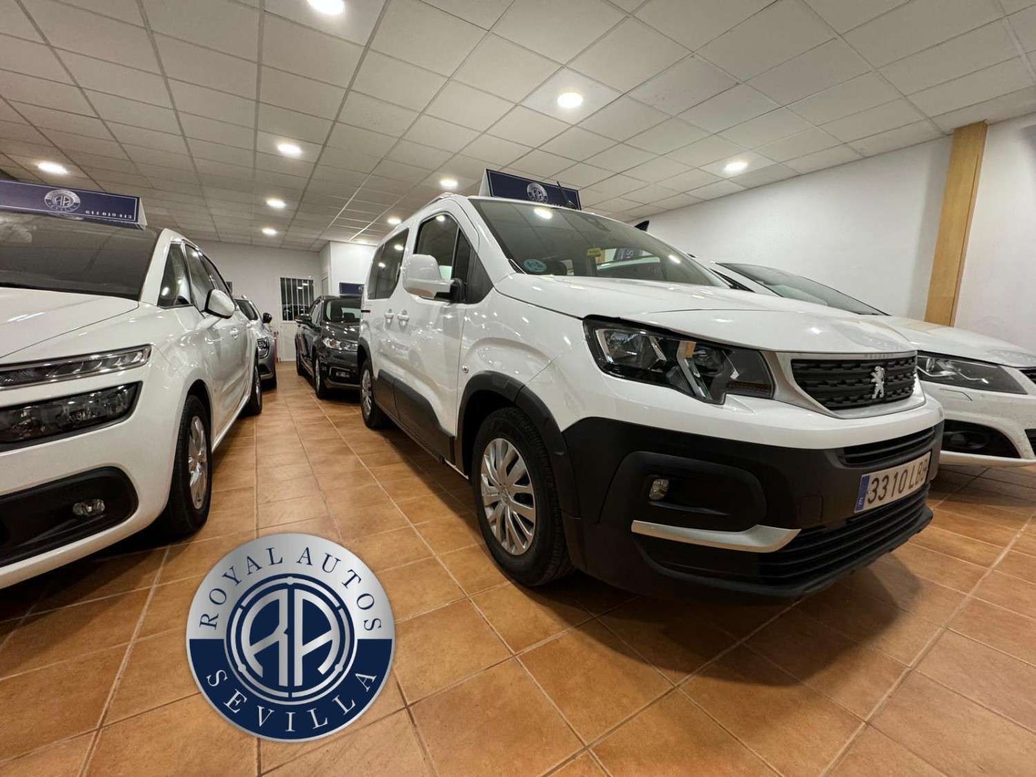 Furgoneta Peugeot blanca en un concesionario, con otros coches al fondo. Logotipo de Royal Autos Sevilla en primer plano.