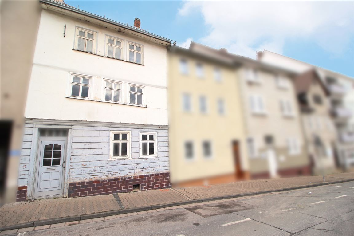 Whitewashed house with peeling paint, red brick base, and multiple windows, next to blurred buildings on a street.