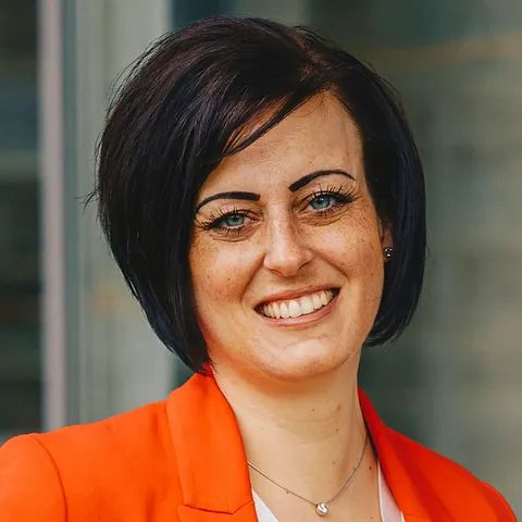 Eine Frau mit einer orangefarbenen Jacke und einer Halskette lächelt in die Kamera.