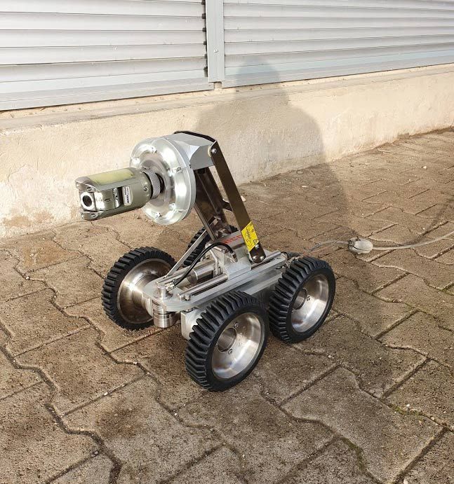 Un petit robot d'inspection argenté à quatre roues, équipé d'une caméra sur un bras, positionné sur une surface en briques.