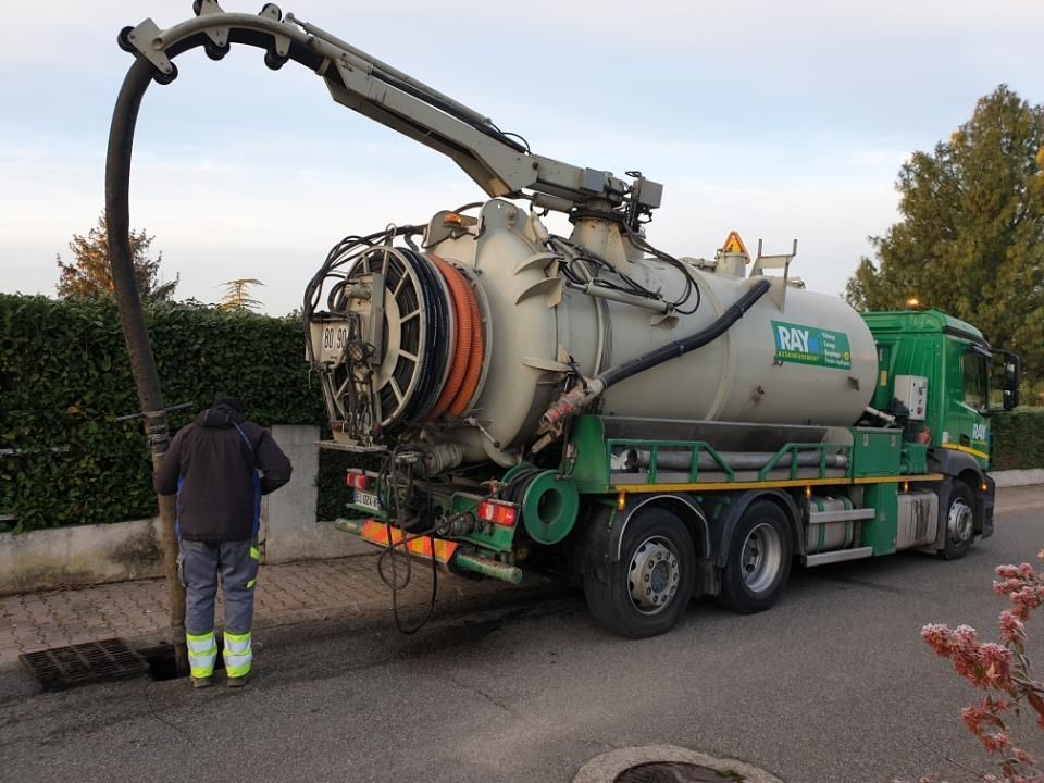 Camion-citerne d'égout avec un tuyau dans une canalisation ; un employé en veste noire et gilet de sécurité se tient à proximité.