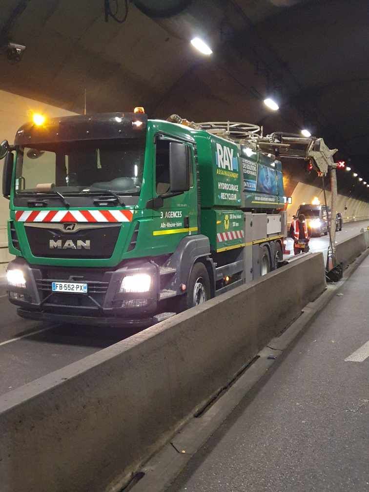 Camion vert dans un tunnel, équipé de matériel d'entretien. Feux allumés, barrière à côté, un autre véhicule en arrière-plan.