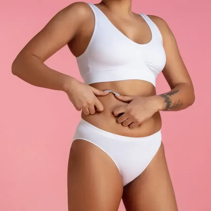Mujer en ropa interior blanca y top corto pellizcando la grasa del vientre, fondo rosa.