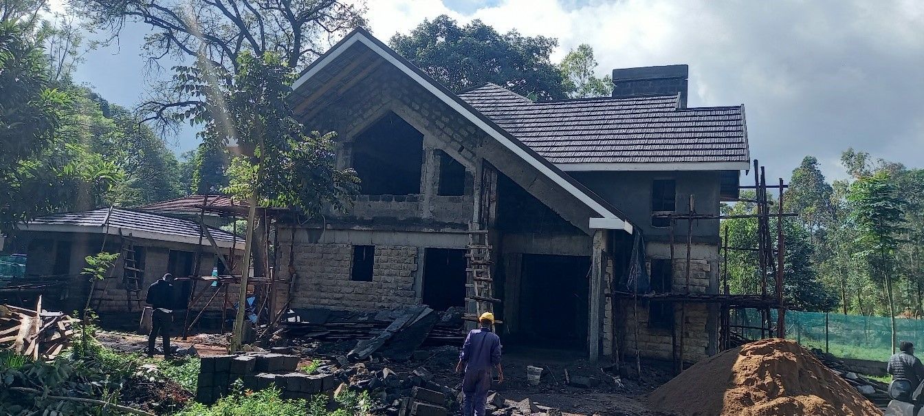 Stone house under construction amid trees, with workers and construction materials visible.