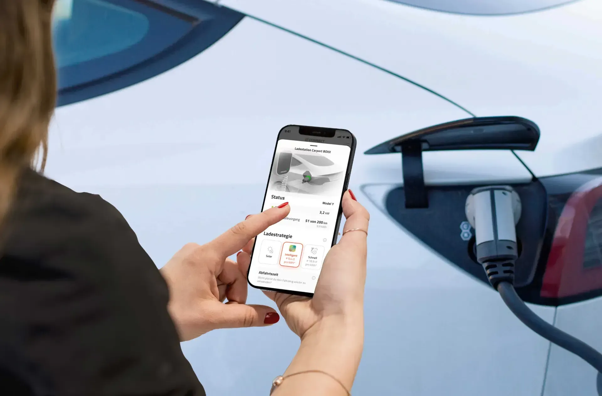 Eine Frau hält ein Mobiltelefon vor ein Elektroauto, das gerade aufgeladen wird.