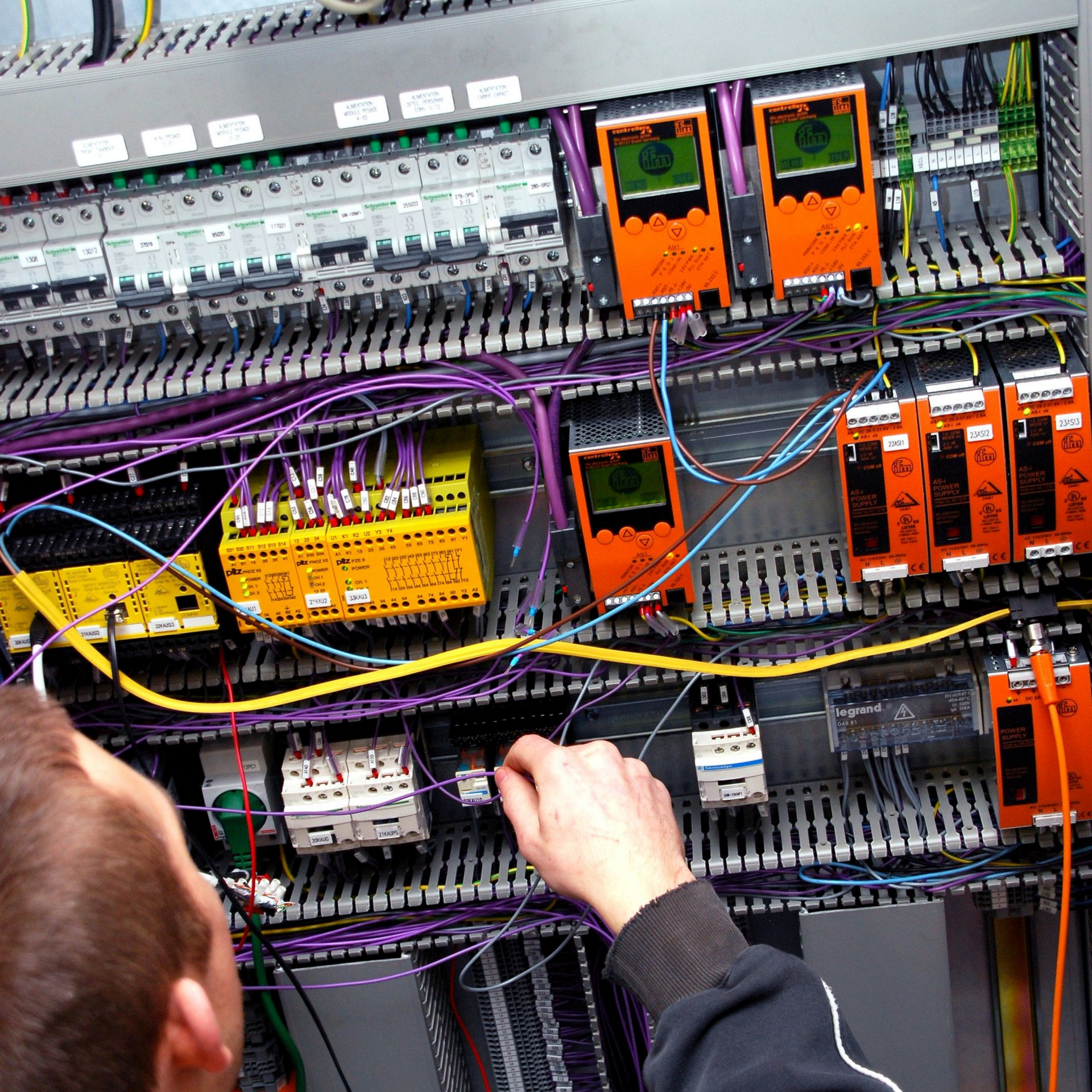 Un électricien travaille sur un tableau électrique ouvert, avec des câblages colorés et des composants modulaires.