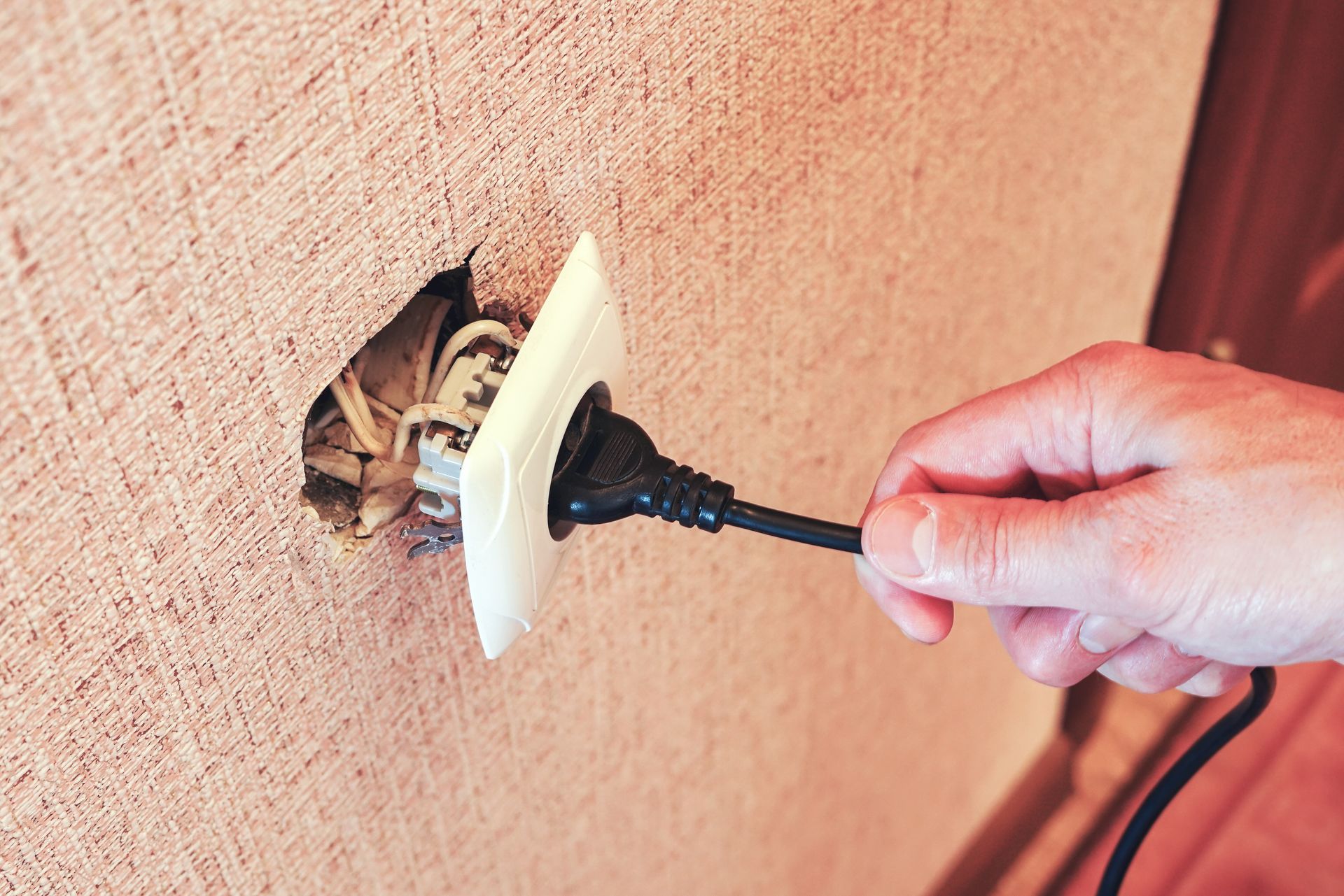 Une main tenant une prise électrique près d'une prise murale détachée et apparente, avec des fils visibles.
