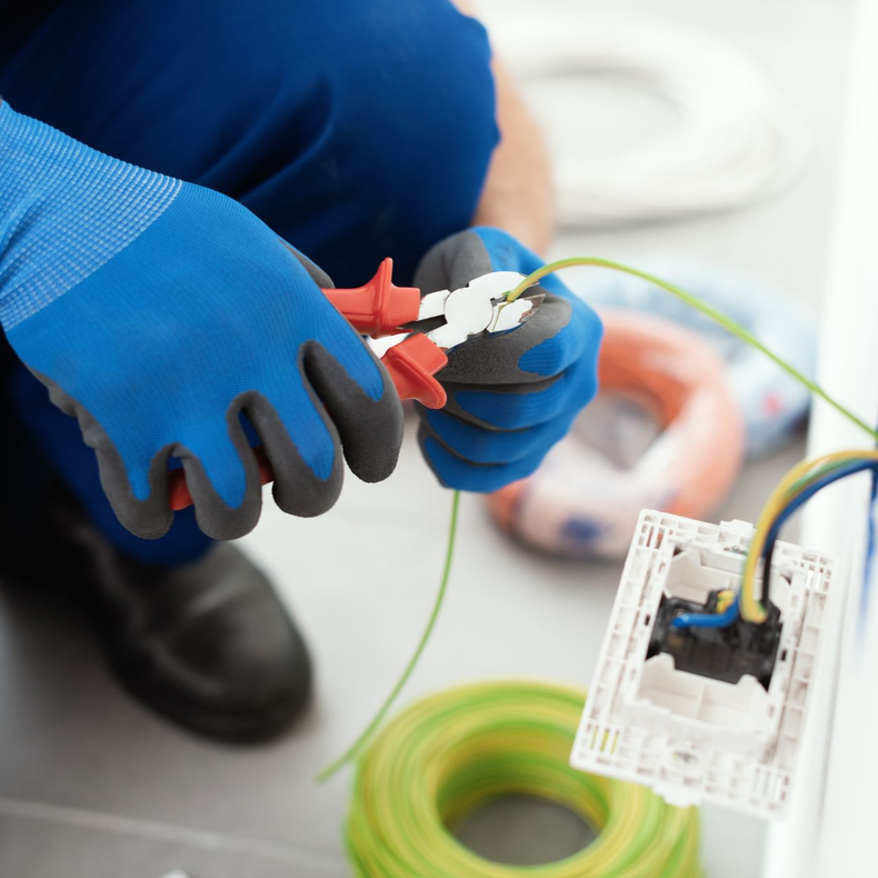 Un électricien portant des gants de protection bleus utilise une pince coupante sur un fil électrique vert près d'une prise murale ouverte.