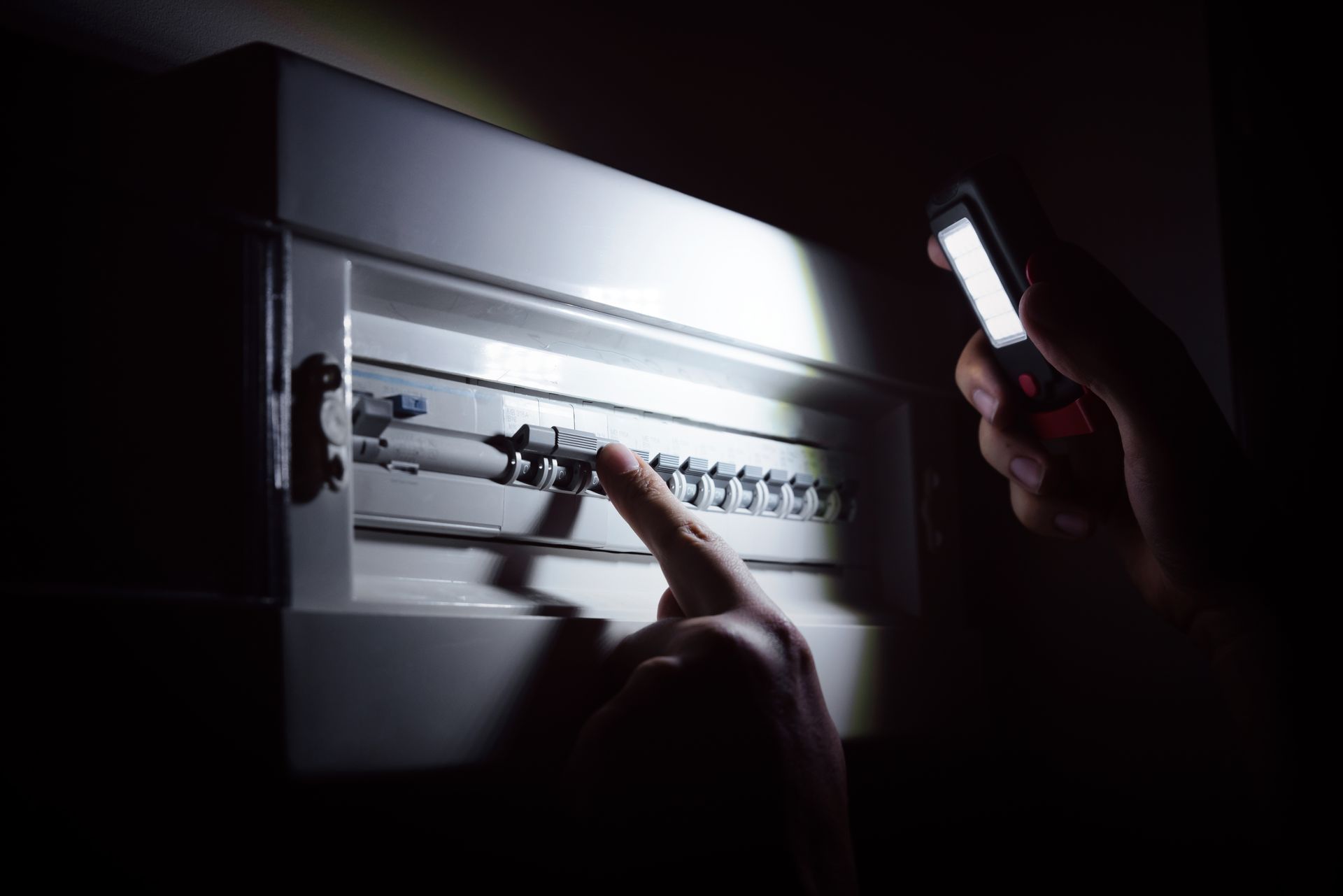 Une personne tient une petite lampe de poche tout en inspectant les interrupteurs d'un tableau électrique dans une pièce sombre.