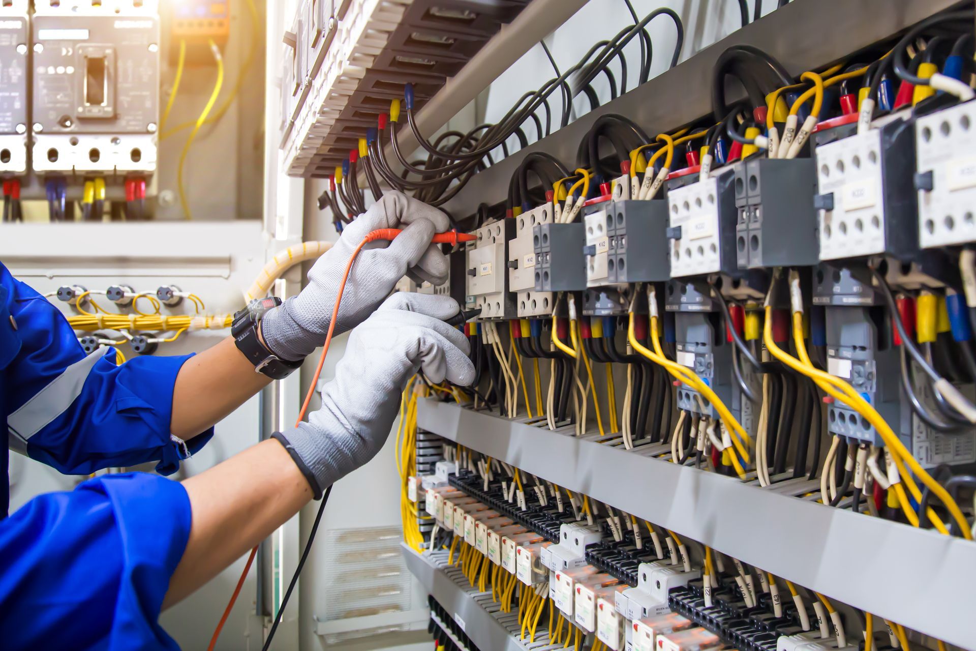 Un technicien en tenue de travail bleue et gants gris utilise un multimètre pour tester le câblage à l'intérieur d'une armoire électrique industrielle.