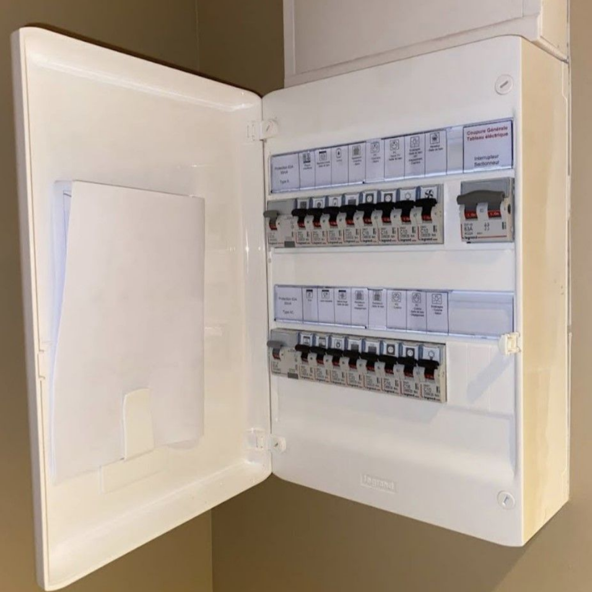Un tableau électrique blanc fixé au mur, porte ouverte, laissant apparaître deux rangées de disjoncteurs.