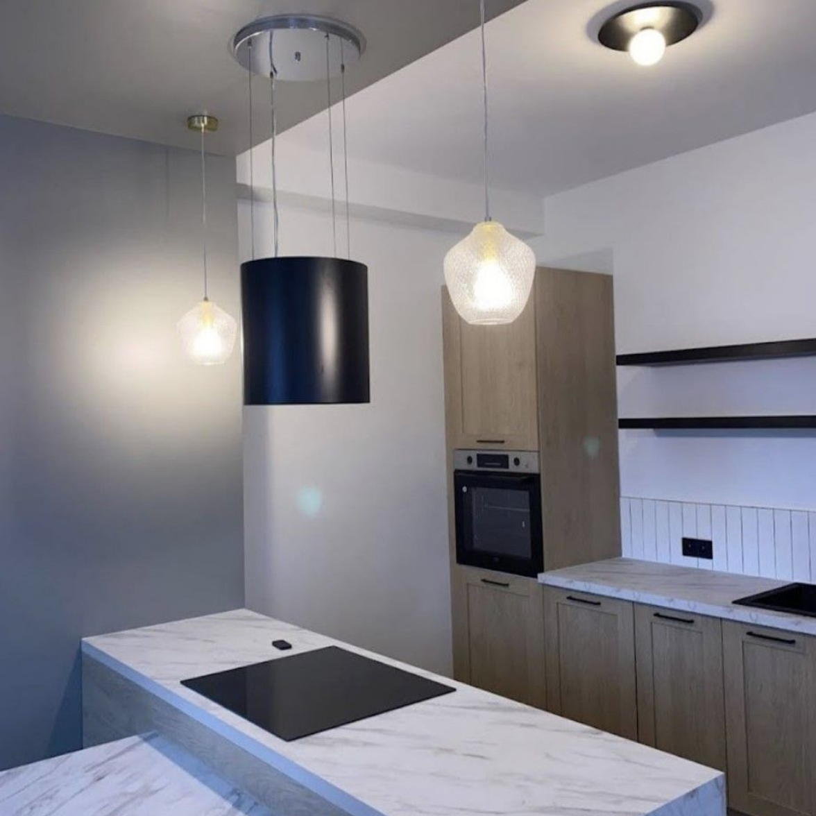 Une cuisine moderne avec un îlot central en marbre, des placards en bois, une hotte noire et des suspensions lumineuses.