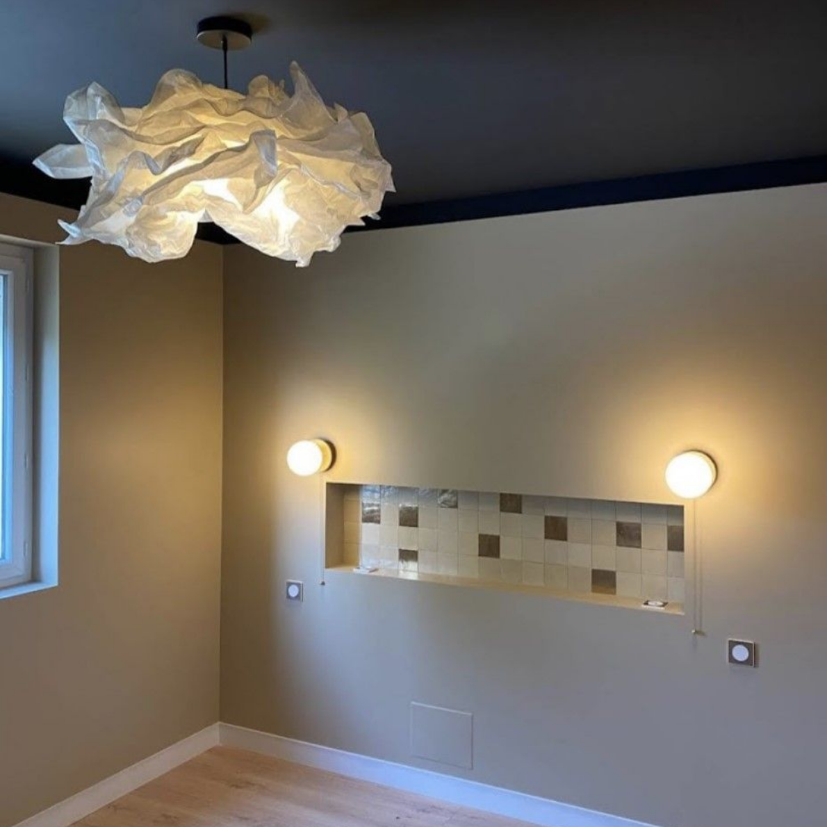 Une pièce avec parquet clair, murs beiges, plafond foncé, suspension blanche texturée et niche murale carrelée.