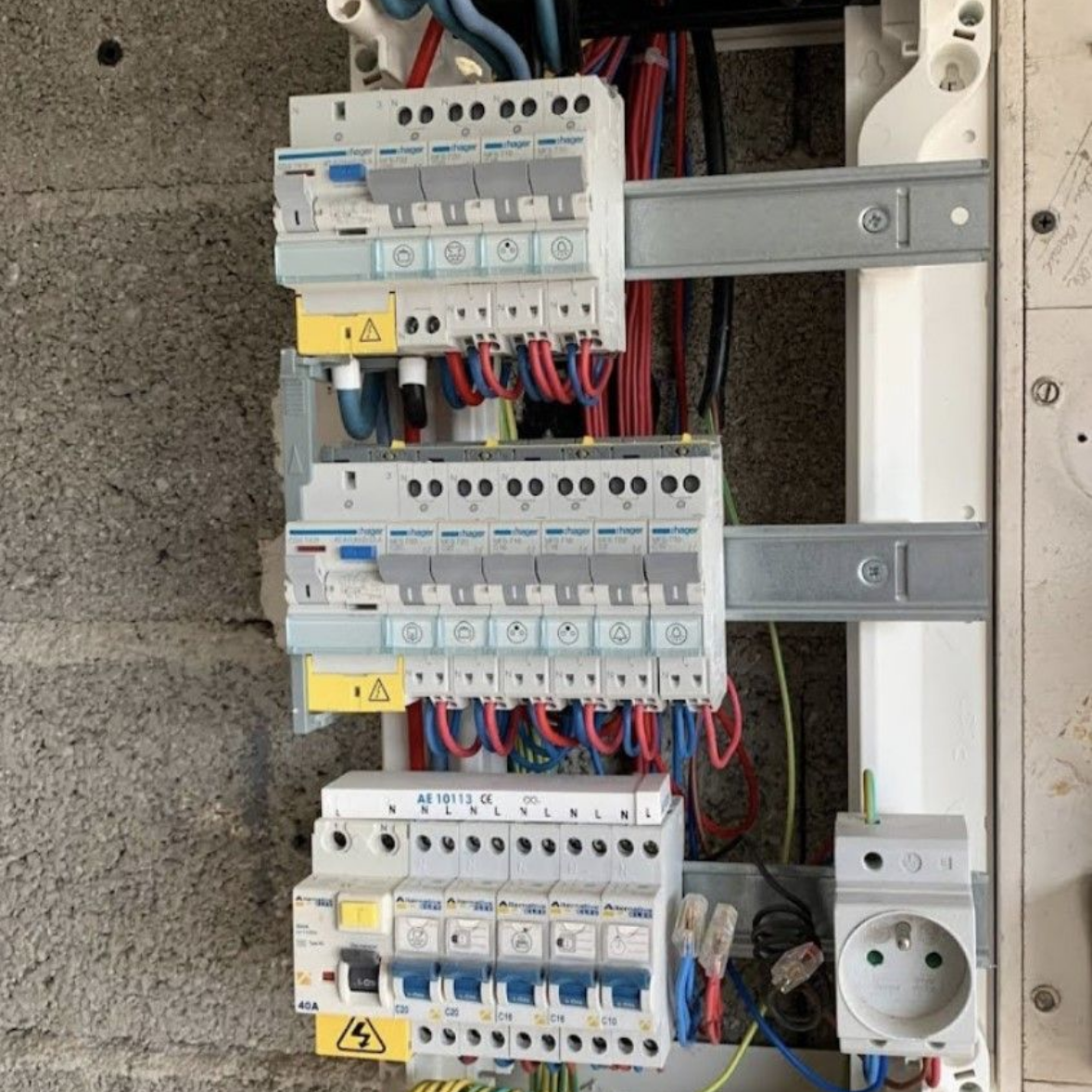 Un tableau électrique installé sur un mur en béton gris, comprenant plusieurs disjoncteurs et une prise de courant.
