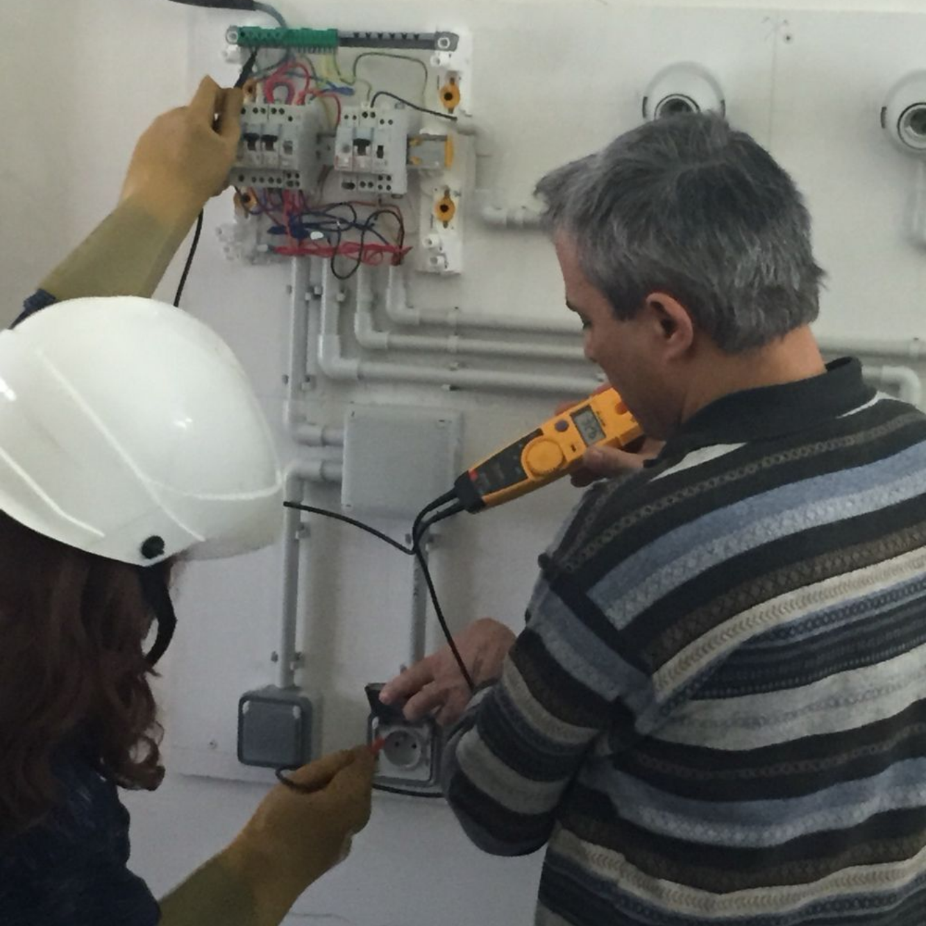Deux techniciens utilisent un multimètre pour tester un panneau électrique mural et une prise de courant.