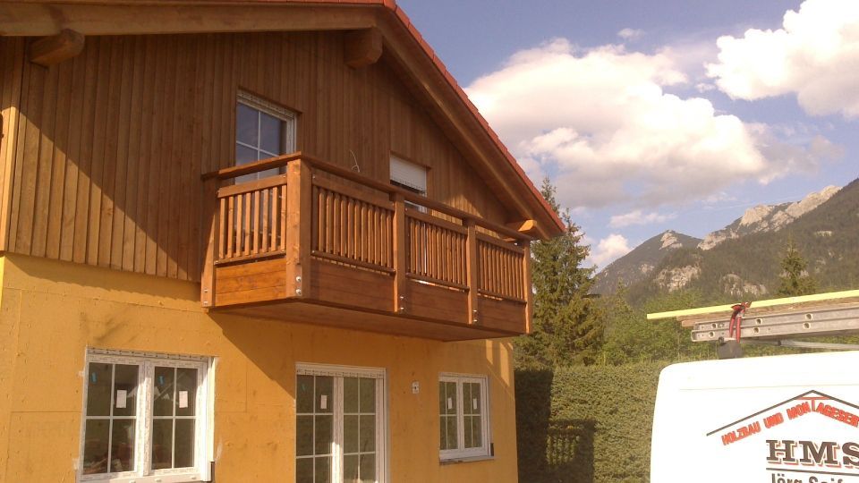 Zweistöckiges beiges Haus mit im Bau befindlichem Balkon an einem sonnigen Tag, mit blauem Himmel und Nachbarhäusern.