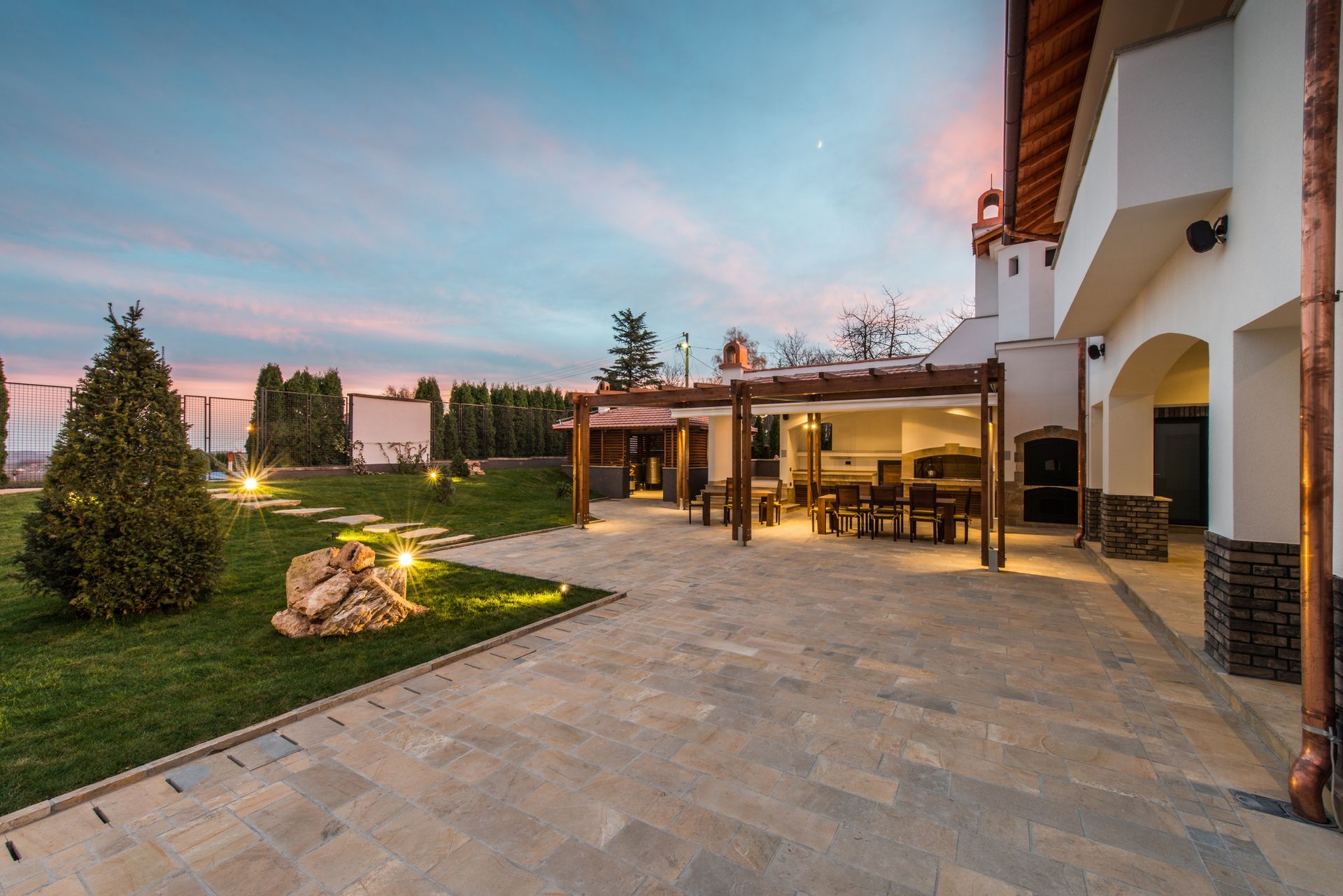 Aménagement extérieur d'une maison avec terrasse en pierre, pergola, éclairage paysager et espaces verts soignés.