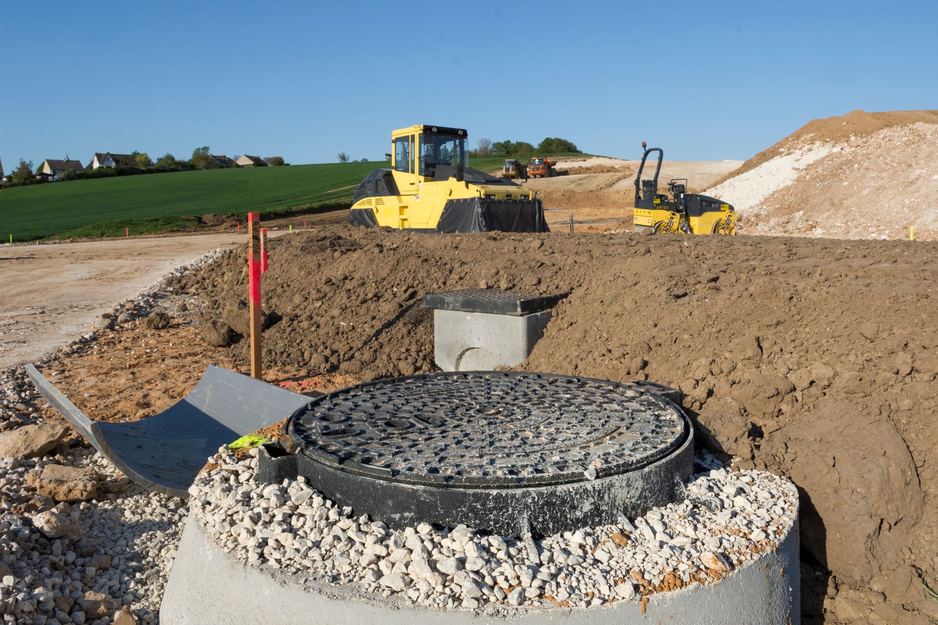 Chantier de construction avec une plaque d'égout en béton dans une tranchée et des engins de chantier en arrière-plan.