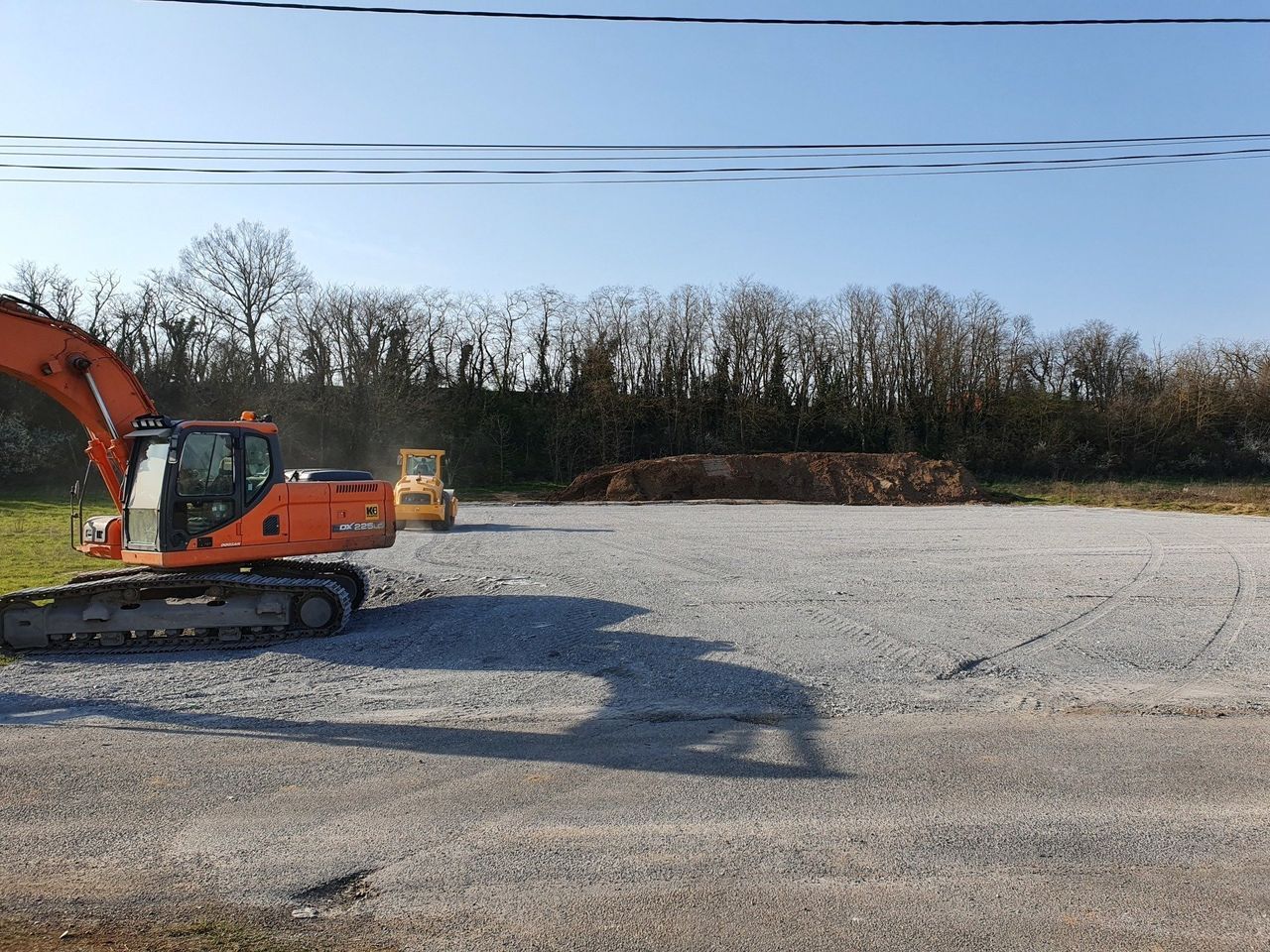 Une pelleteuse orange garée sur un terrain gravillonné, bordée d'arbres.