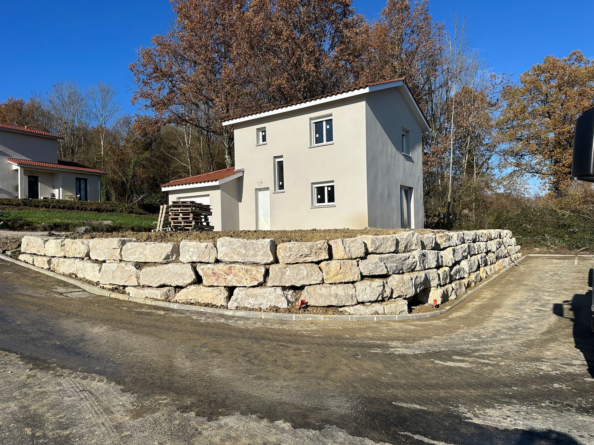 Vue sur un muret de soutènement en pierre, avec une maison blanche à deux étages fraîchement construite.