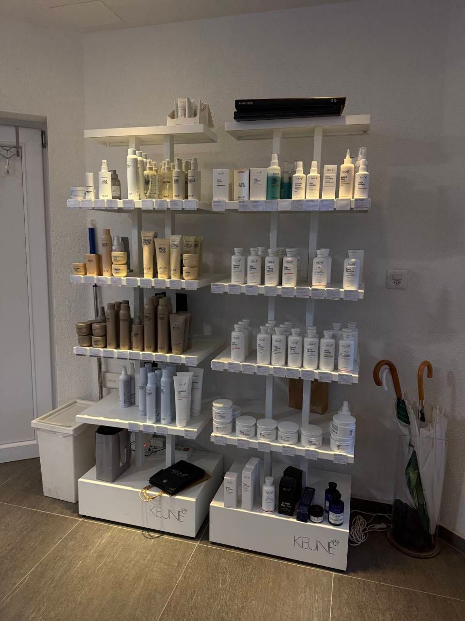 Weisses Regal mit Kosmetikprodukten in einem Ladenambiente, daneben Regenschirme und ein Abfalleimer.